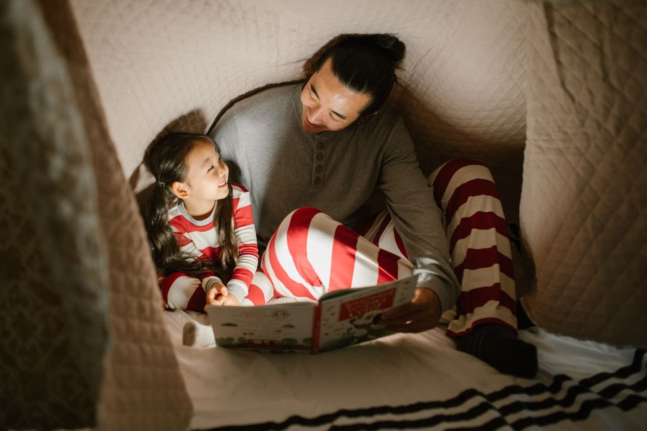 Father and daughter enjoy a bedtime story under a cozy blanket, sharing smiles and bonding.