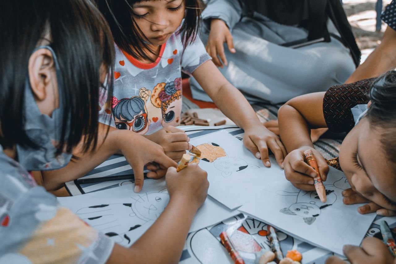 Crop diverse focused group of little children sitting at table and learning to draw cat on paper sheets with crayon pencils in kindergarten in daylight